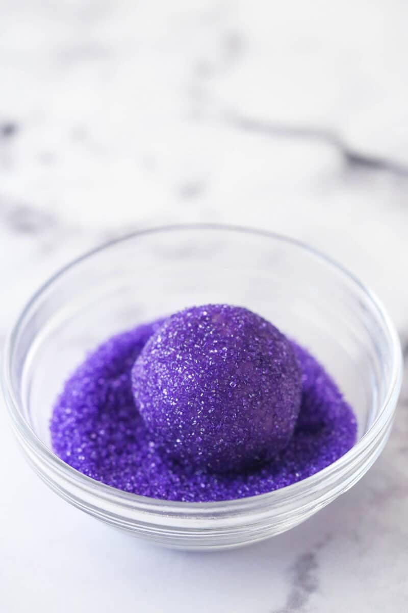 A ball of purple cookie dough being coated in purple sanding sugar for Halloween Monster Cookies.