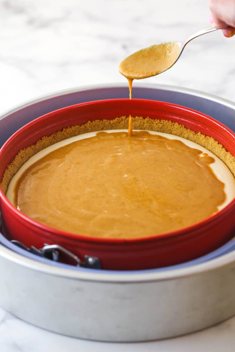 Spooning pumpkin pie filling over vanilla cheesecake filling in a springform pan in a water bath.