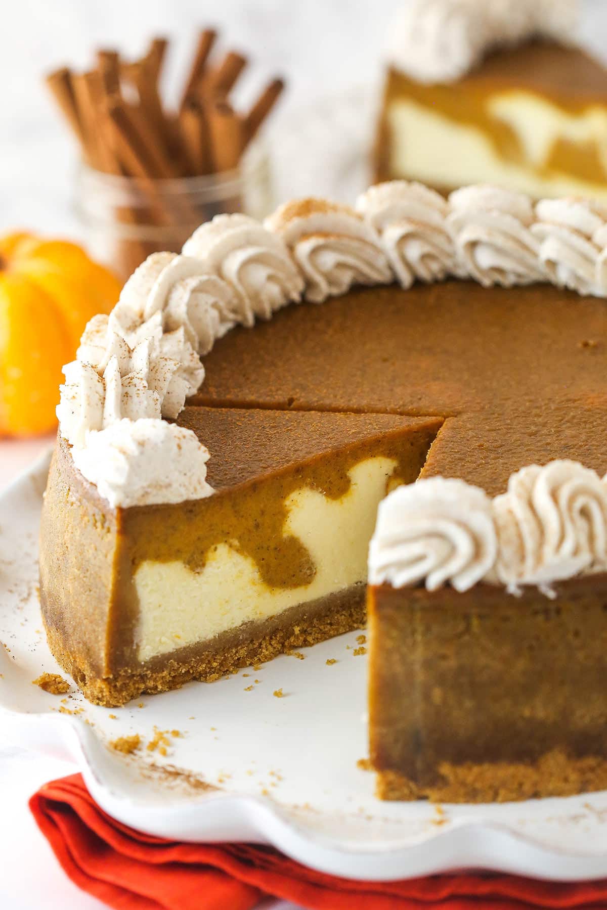Pumpkin pie cheesecake is made of graham cracker crust topped with vanilla cheesecake, pumpkin pie filling, and fluffy whipped cream. Pumpkin pie cheesecake with a slice taken out of it.