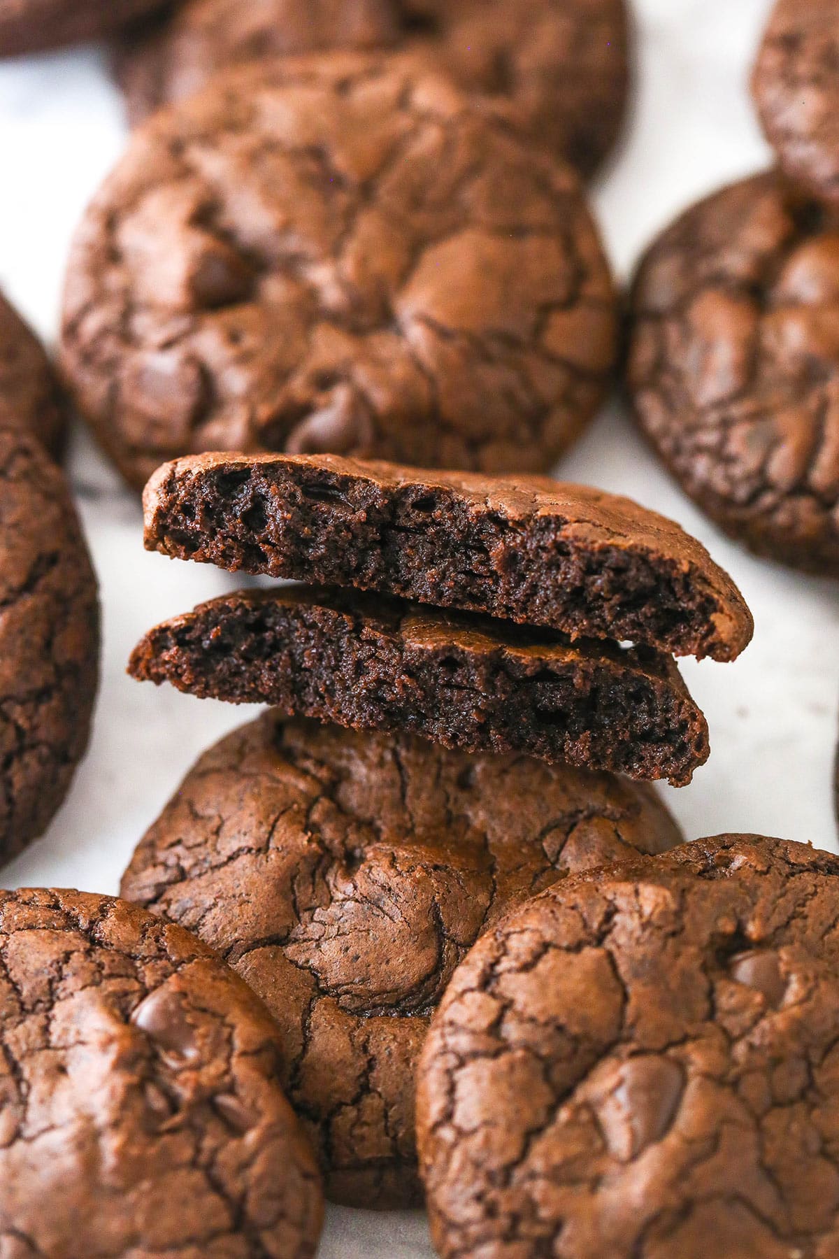Chewy, dense chocolate cookies with the crackly top of a brownie and chocolate chips scattered throughout, these brownie cookies have it all. Brownie cookies scattered on a marble surface. One is broken in half.