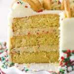 Frosted Sugar Cookie Layer Cake on a white cake plate with several slices removed to show layers.