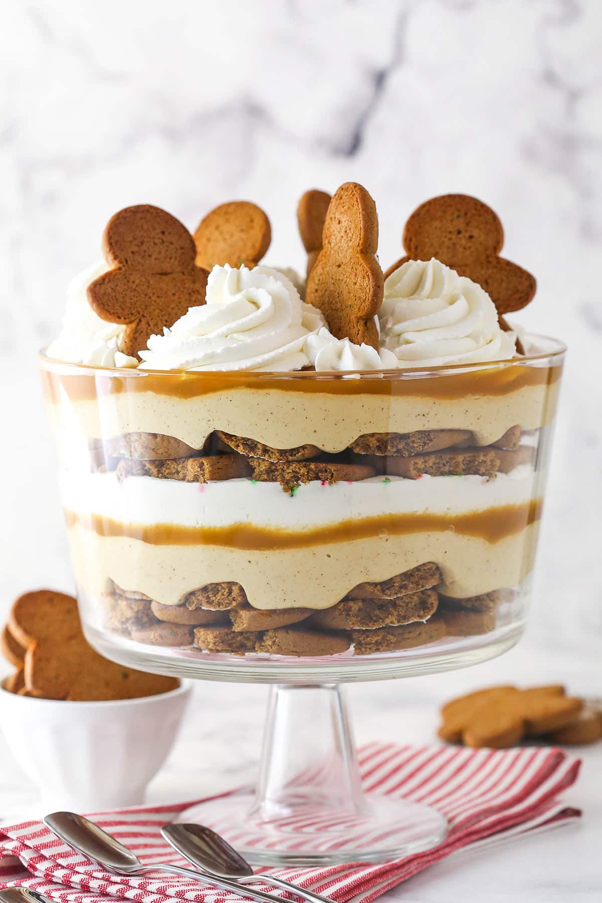 This Gingerbread Cheesecake Trifle is an easy no-bake holiday dessert! It has layers of thick gingerbread cookies, no-bake gingerbread cheesecake, caramel and whipped cream. A Gingerbread Cheesecake Trifle in a trifle bowl on a striped napkin.