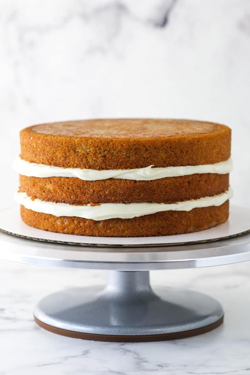 3 layers of Spice Cake with frosting in between them on a silver cake stand and cardboard round.