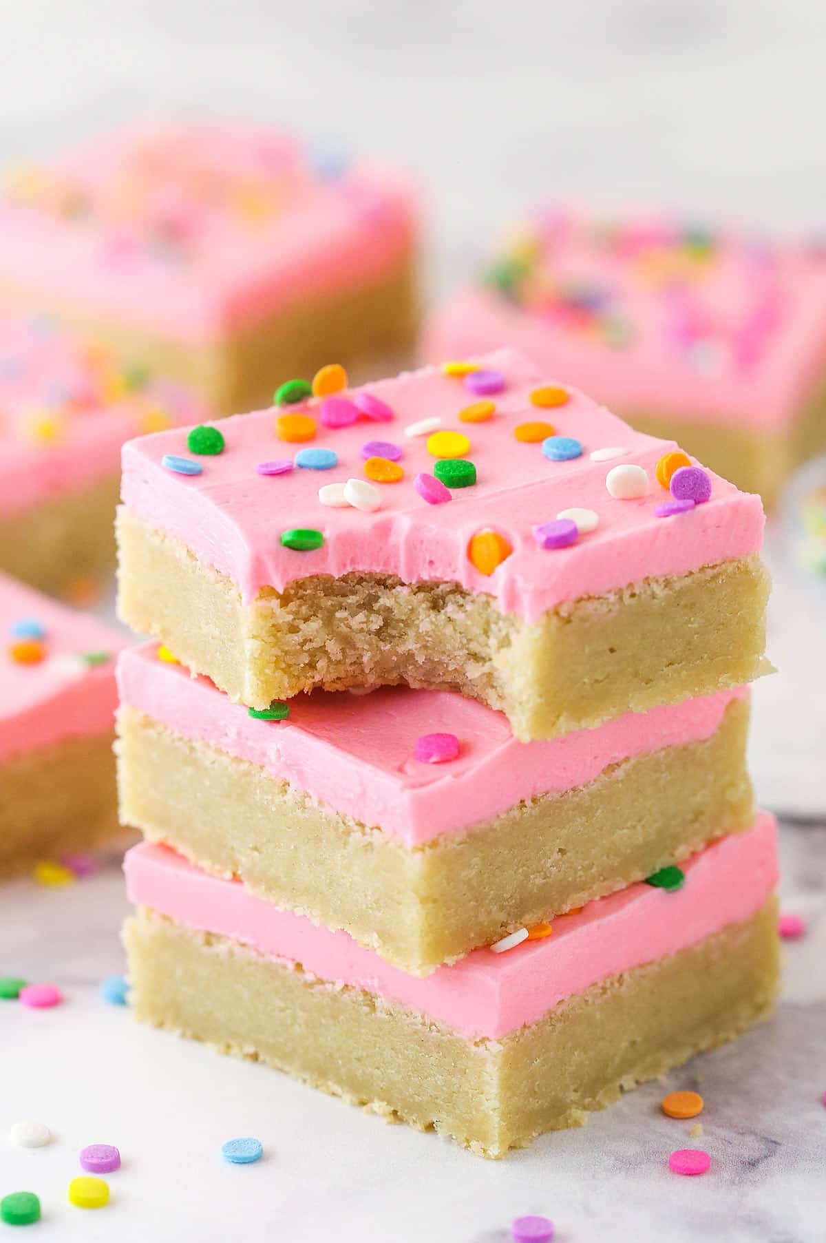 Soft, chewy, and full of sweet vanilla flavor, these easy frosted sugar cookie bars will become a new dessert table favorite. A stack of sugar cookie bars. The top one has a bite taken out of it.