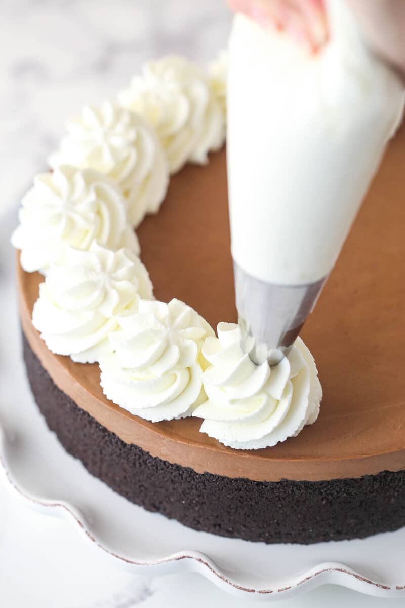 Hand using a piping bag to pipe whipped cream around the outside of a No-Bake Chocolate Cheesecake.