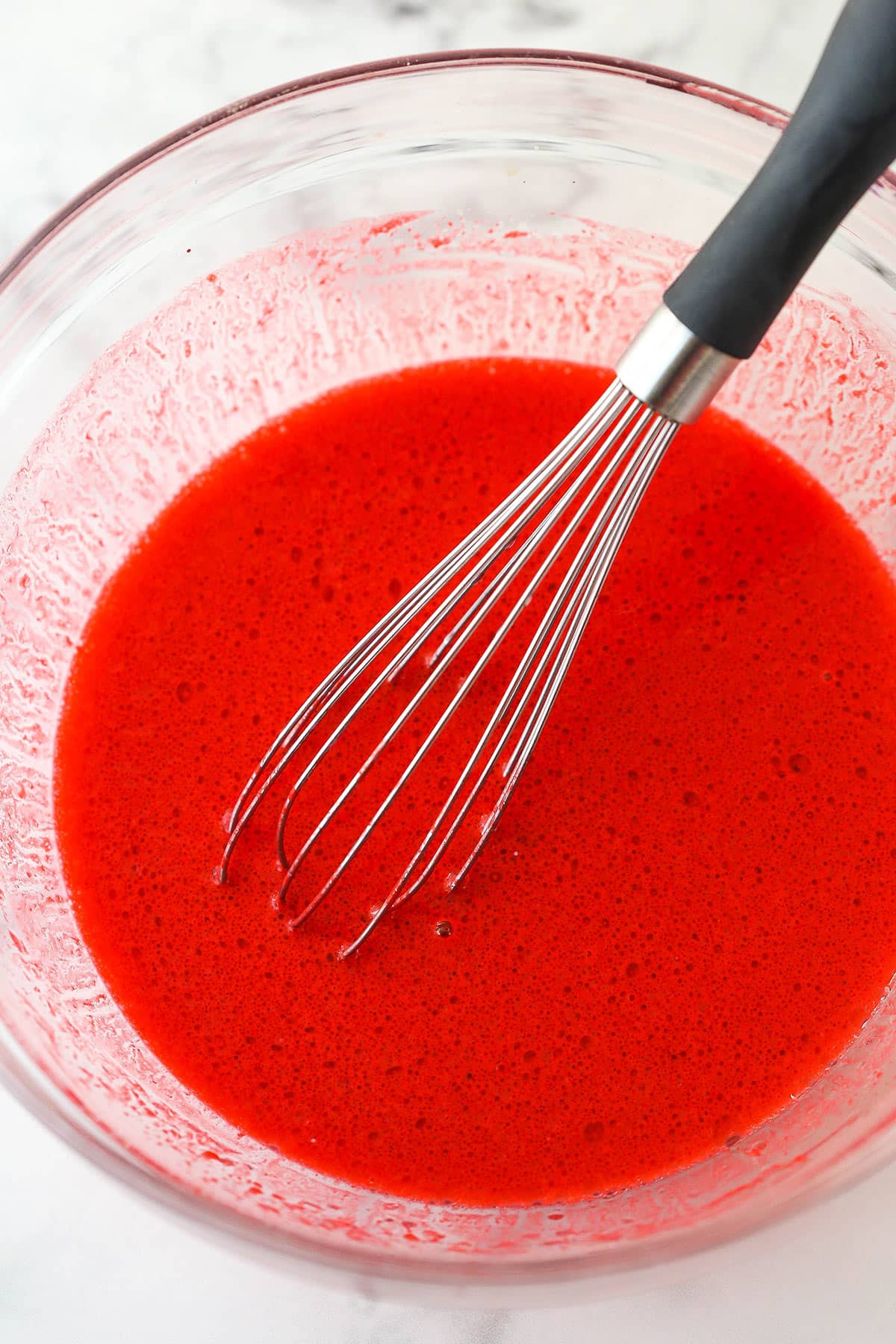 Whisking together buttermilk, vegetable oil, vanilla extract, eggs, vinegar, and food coloring for cupcake batter.