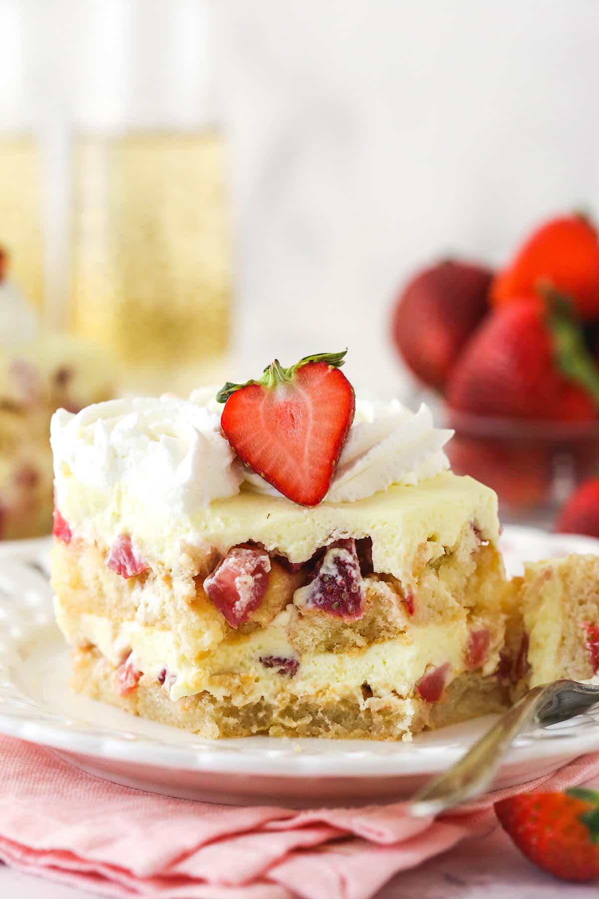 Strawberry Champagne Tiramisu is such a fun and tasty twist on classic tiramisu. Ladyfingers are dipped in a lightly-sweet champagne mixture and layered with chopped strawberries and delicious mascarpone filling. A slice of Strawberry Champagne Tiramisu on a small white plate with a bite removed to show layers.
