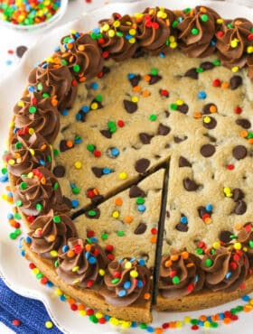 A full Chocolate Chip Cookie Cake with a slice cut out on a white platter.