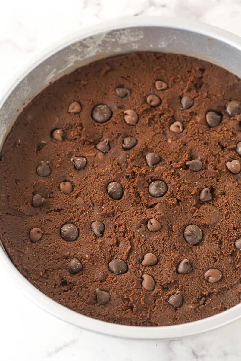 Triple chocolate cookie cake is made up of a fudgy chocolate cookie base loaded with chocolate chips and topped with chocolate buttercream. Chocolate cookie dough in a cake pan sprinkled with chocolate chips.