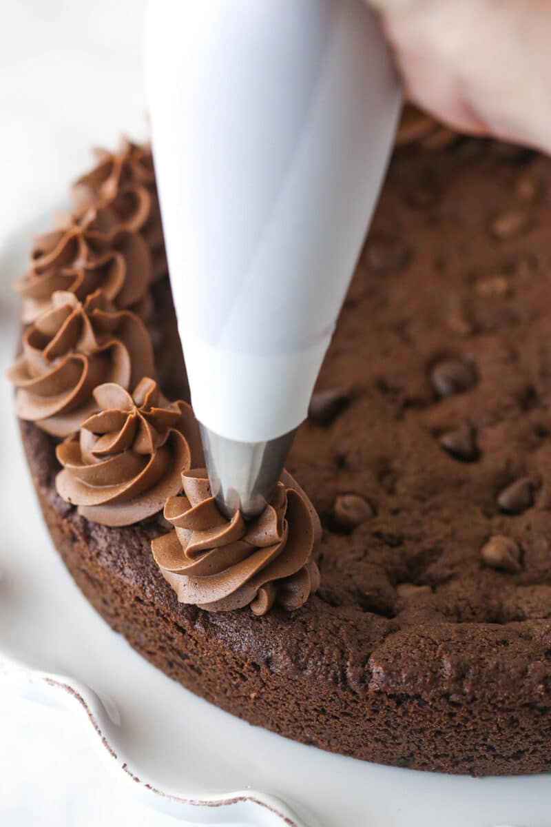 Triple chocolate cookie cake is made up of a fudgy chocolate cookie base loaded with chocolate chips and topped with chocolate buttercream. Piping swirls of chocolate buttercream around the edge of a chocolate cookie cake.