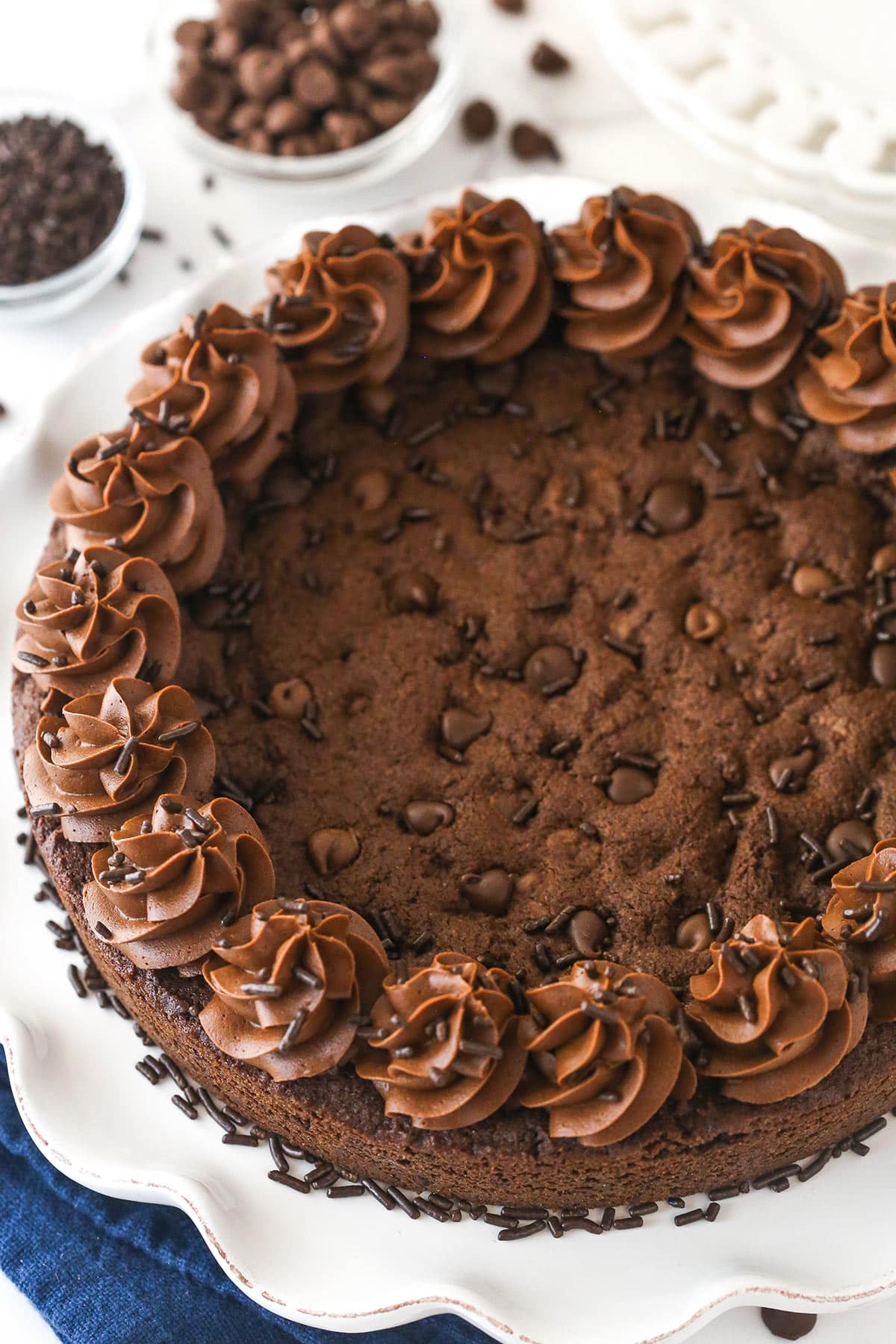 Triple chocolate cookie cake is made up of a fudgy chocolate cookie base loaded with chocolate chips and topped with chocolate buttercream. Overhead image of chocolate cookie cake on a serving platter.