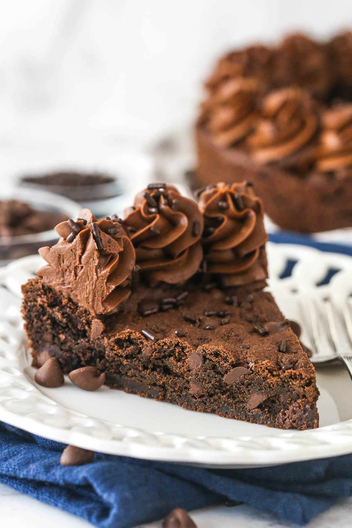Triple chocolate cookie cake is made up of a fudgy chocolate cookie base loaded with chocolate chips and topped with chocolate buttercream. A slice of chocolate cookie cake on a plate.