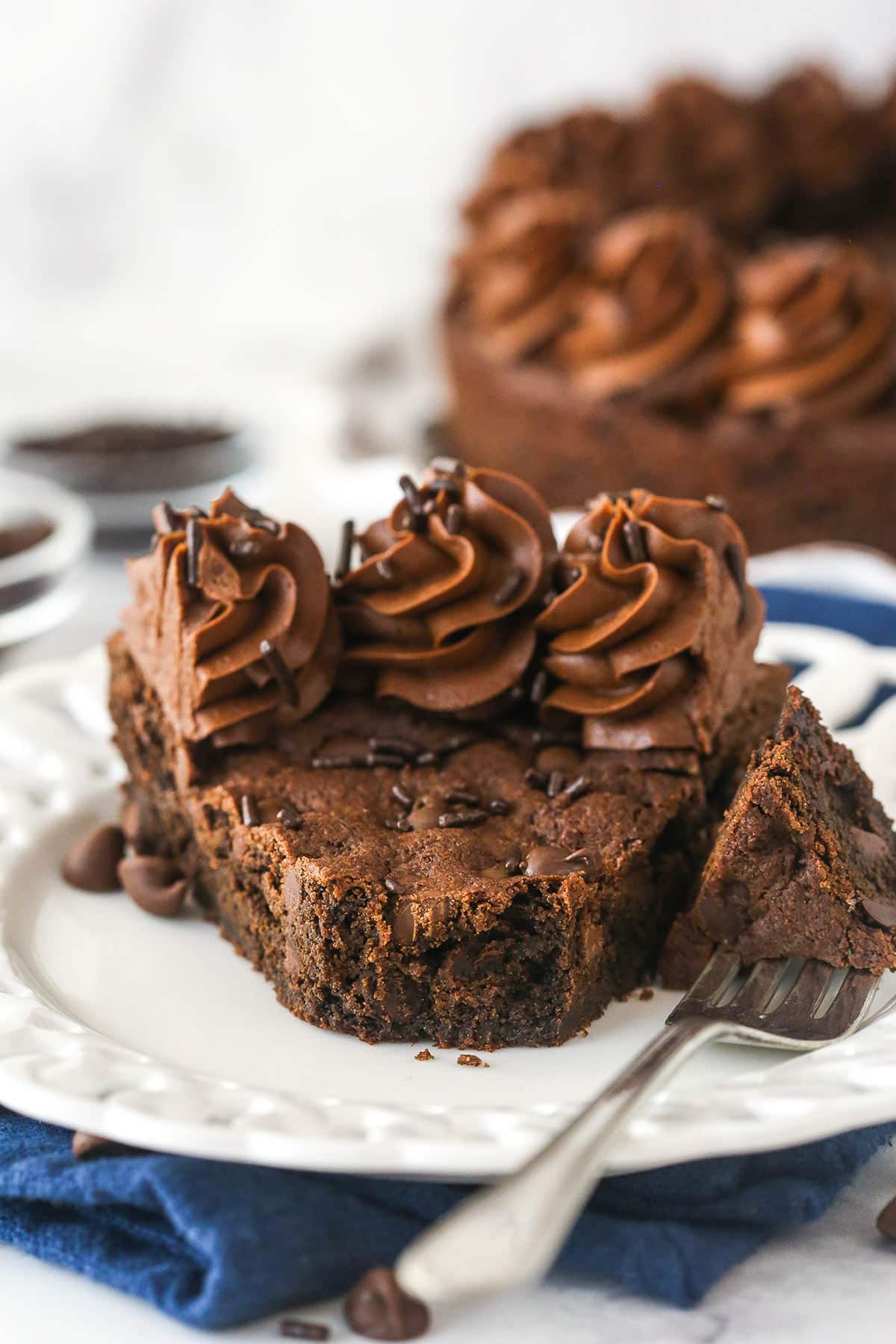 Triple chocolate cookie cake is made up of a fudgy chocolate cookie base loaded with chocolate chips and topped with chocolate buttercream. A fork taking a bite out of a slice of chocolate cookie cake.