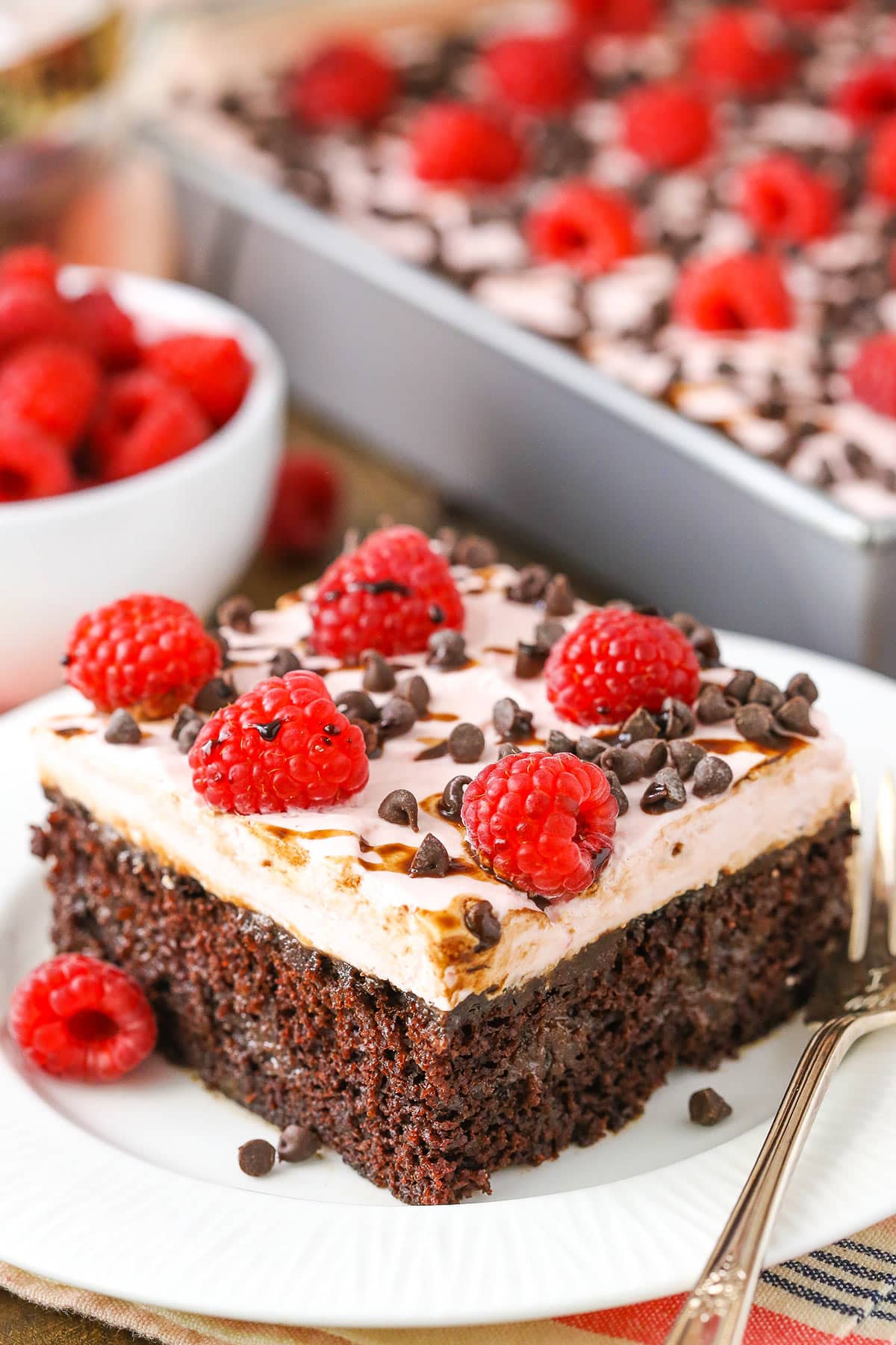 A square serving of Raspberry Chambord Chocolate Poke Cake next to a silver fork on a white plate