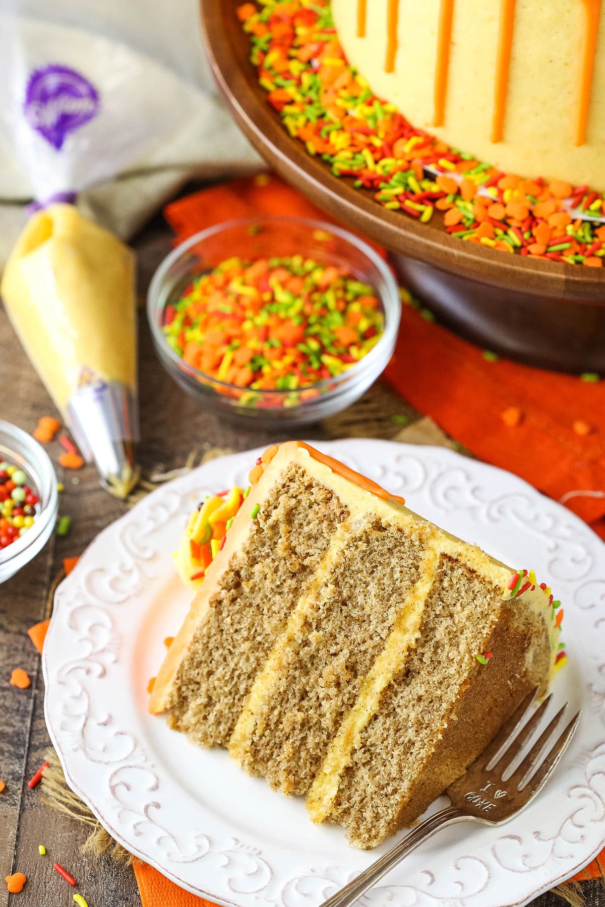 A slice of Spice Cake with Pumpkin Mascarpone Buttercream on a white plate with sprinkles and frosting in the background