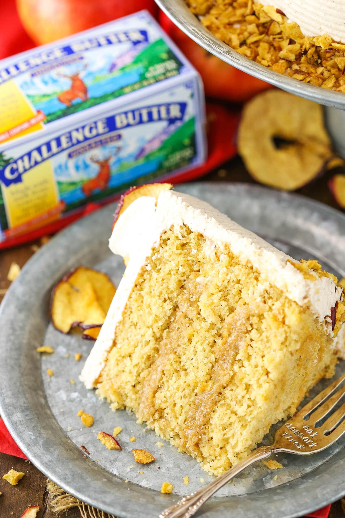 A slice of Cinnamon Apple Layer Cake next to a silver fork on a gray plate with a box of Challenge Butter in the background