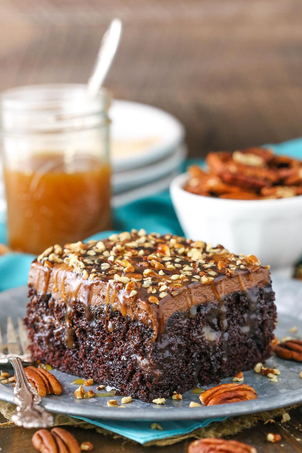 This Chocolate Turtle Poke Cake is a moist chocolate cake soaked with caramel sauce and topped with caramel chocolate frosting and chopped pecans! A square serving of Chocolate Turtle Poke Cake next to a silver fork on a gray plate