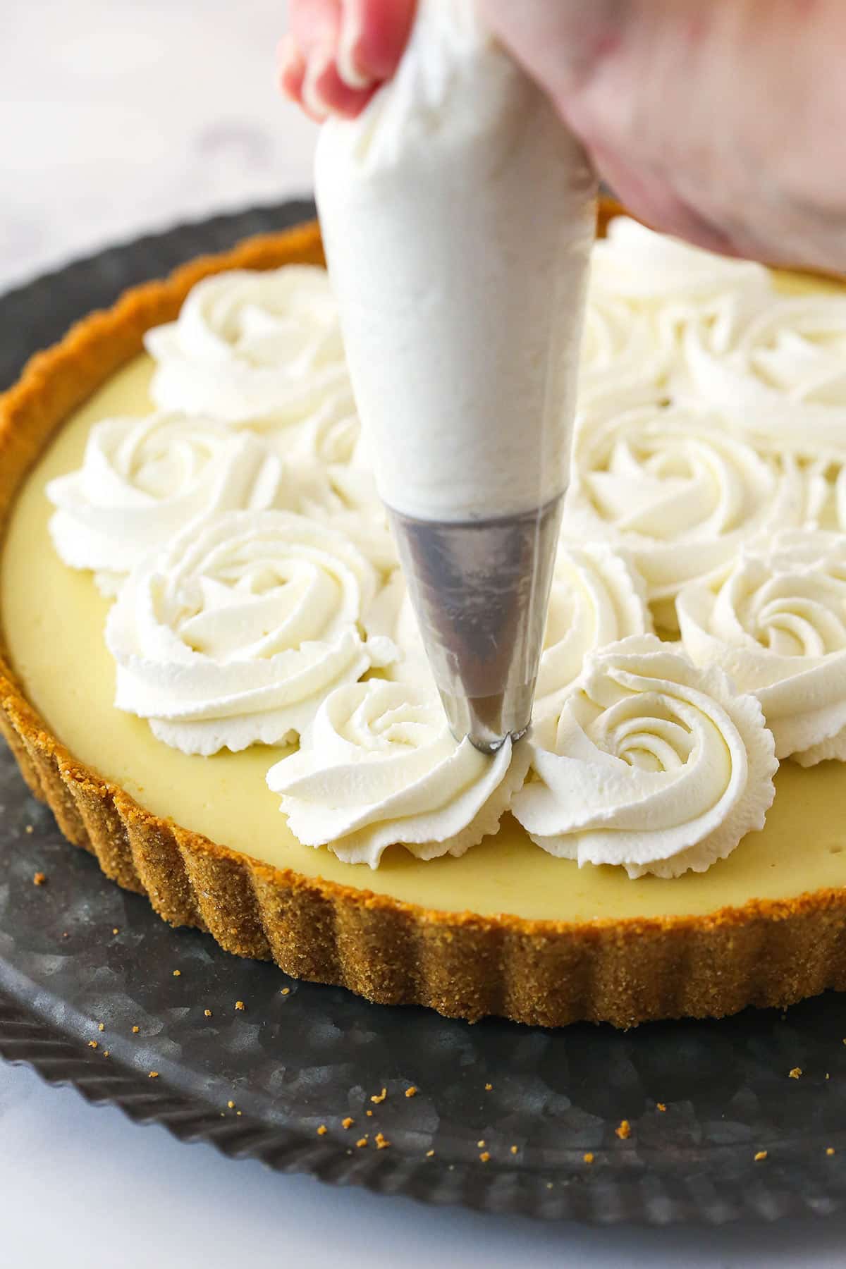 This lemon tart is made up of graham cracker crust, a sweet and tangy, creamy lemon filling, and a fluffy whipped cream topping. Piping whipped cream over the top on a lemon tart.