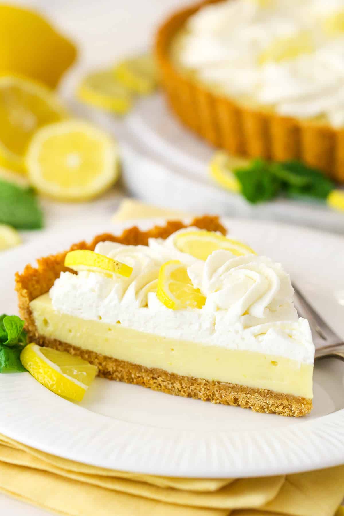 This lemon tart is made up of graham cracker crust, a sweet and tangy, creamy lemon filling, and a fluffy whipped cream topping. A slice of lemon tart on a plate.