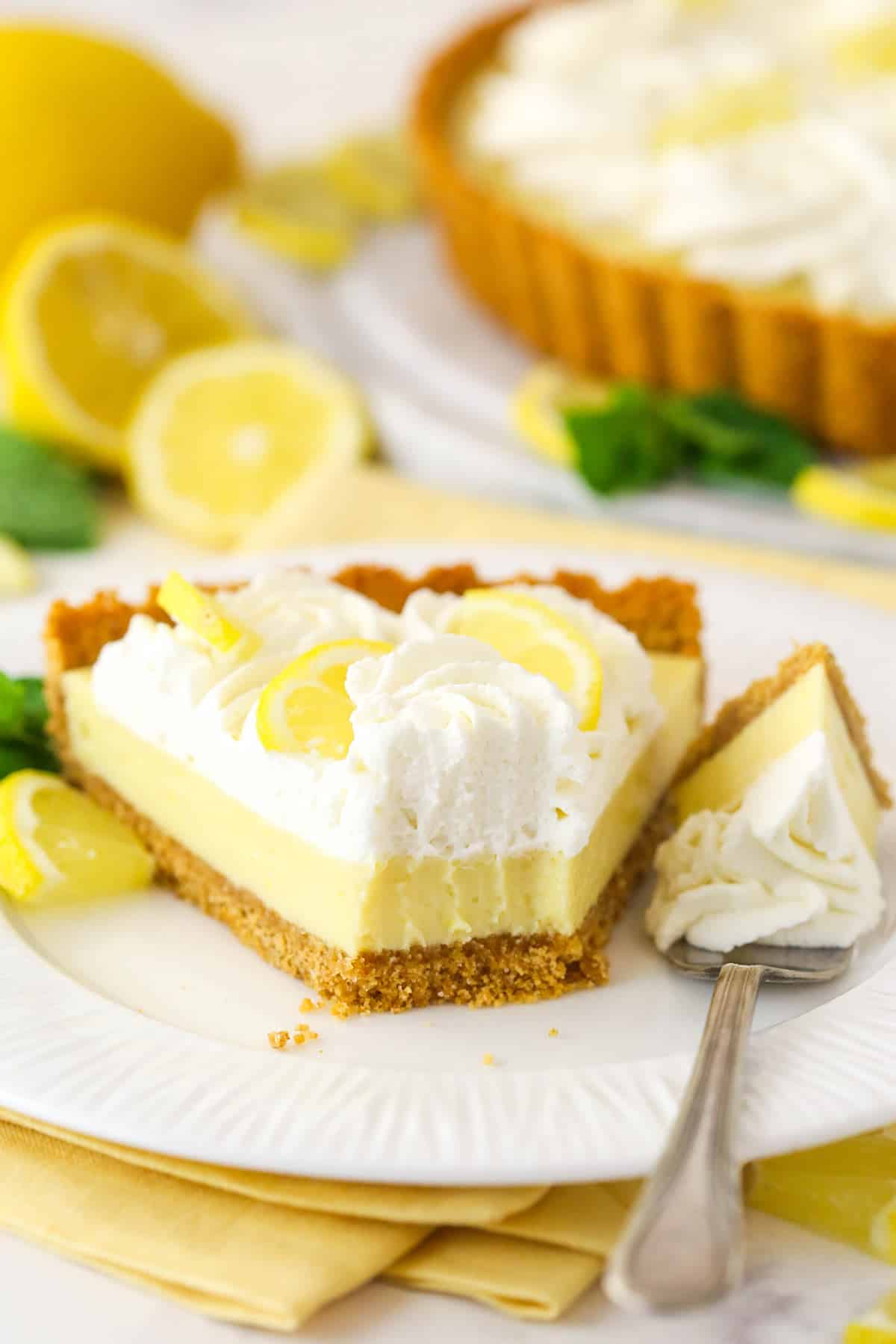 This lemon tart is made up of graham cracker crust, a sweet and tangy, creamy lemon filling, and a fluffy whipped cream topping. A fork taking a bite out of a slice of lemon tart.