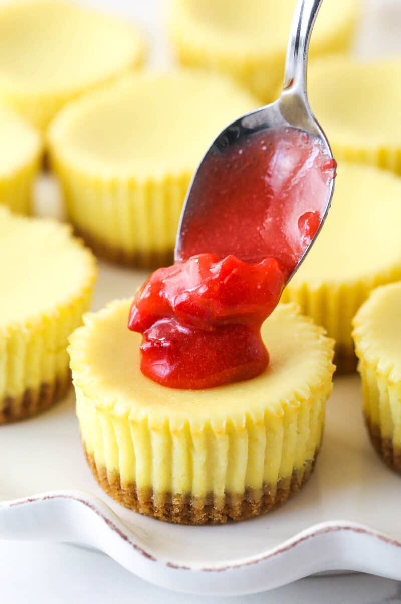 Mini strawberry cheesecakes are made of graham cracker crusts, thick, creamy vanilla cheesecake filling, and sweet strawberry sauce. Spooning strawberry topping onto mini vanilla cheesecakes.