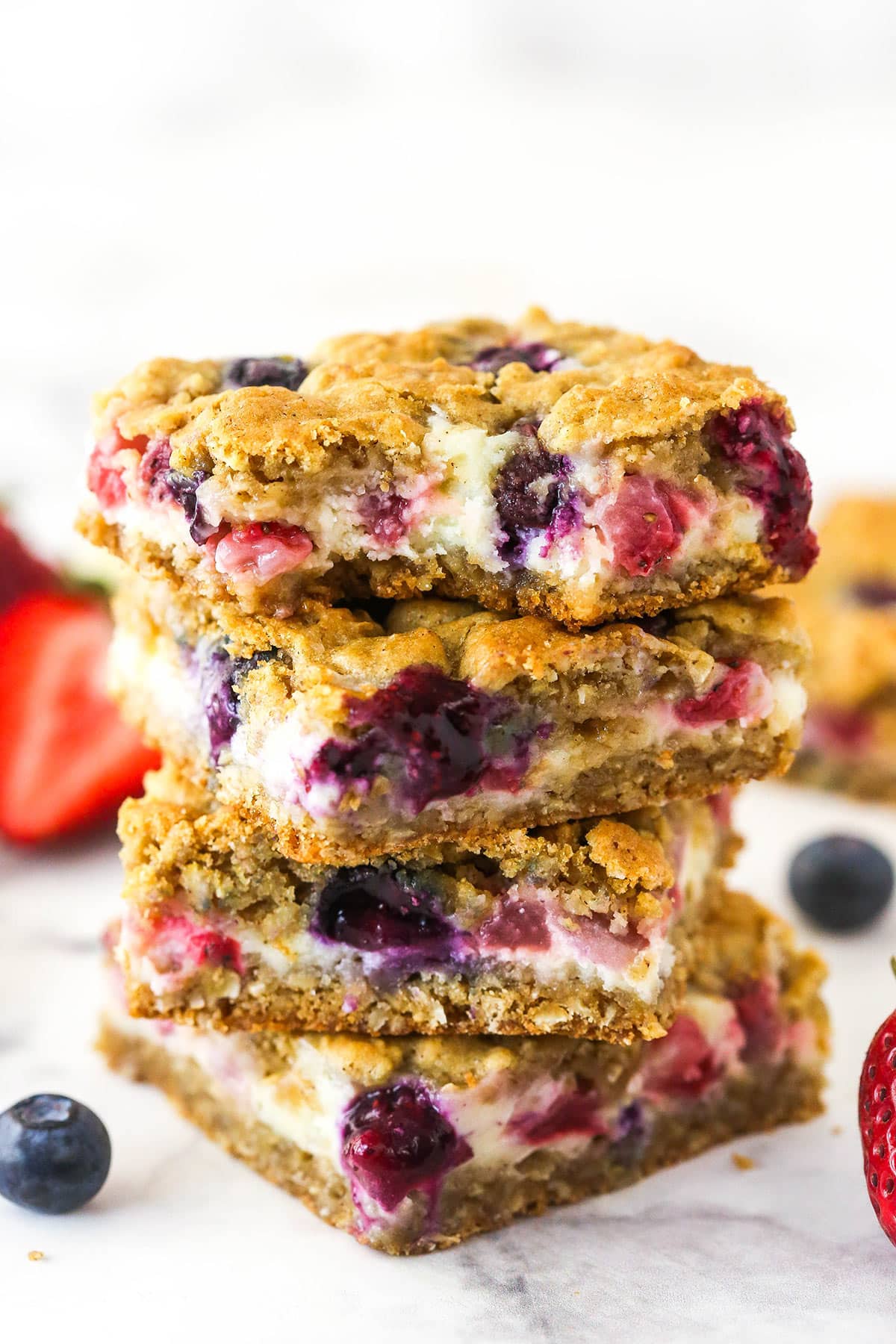 Oatmeal cheesecake bars are made with an oatmeal cookie base topped with a creamy cheesecake layer, lots of berries, and cookie crumbles. A stack of berry oatmeal cheesecake bars. The top one has a bite taken out of it.