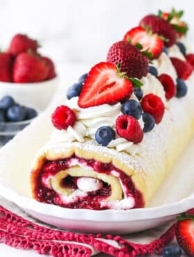 decorated Berry Vanilla Cake Roll on a white platter with berries in the background