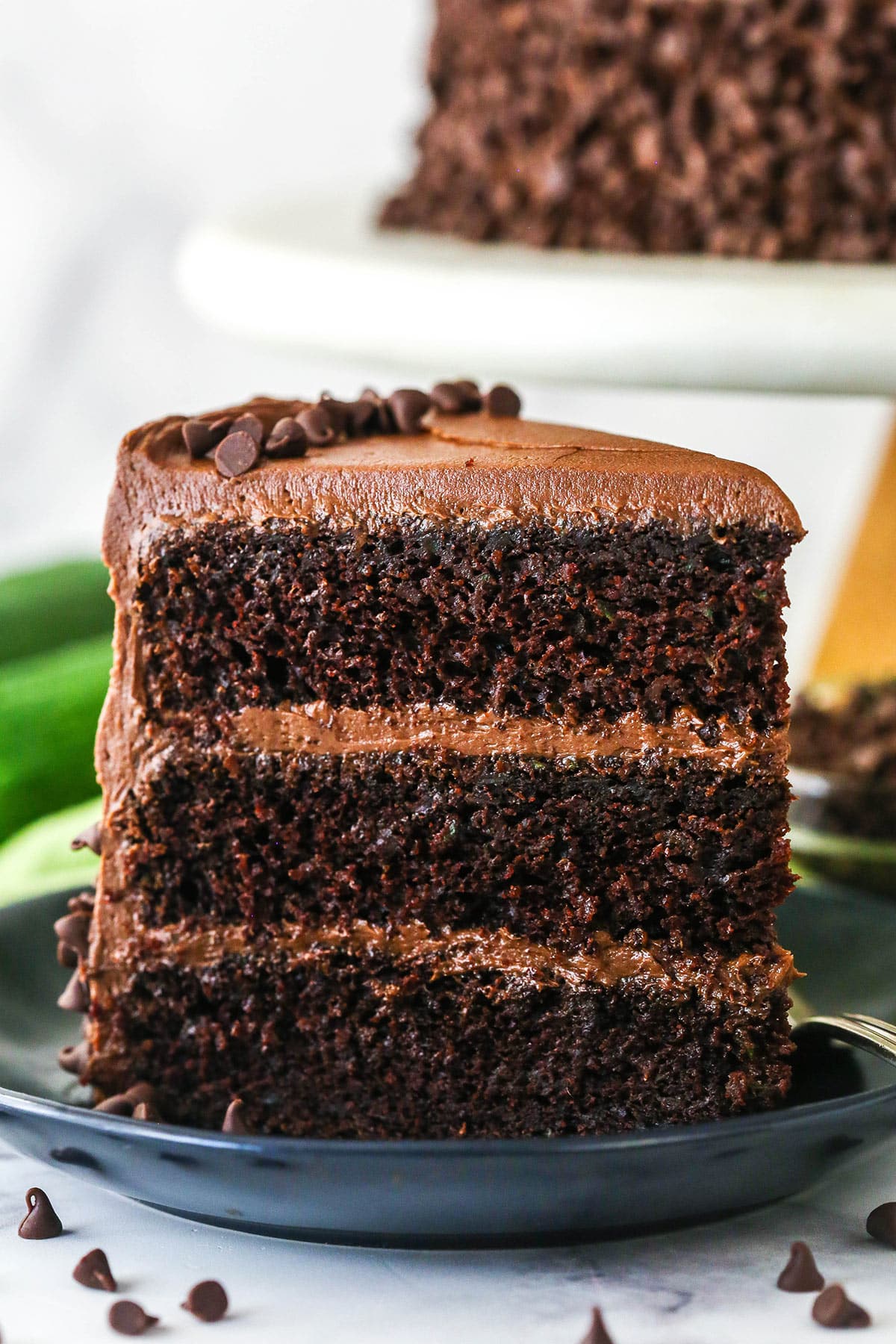 This Chocolate Zucchini Cake is so dreamy! 3 moist, zucchini-loaded chocolate cake layers are filled and frosted with chocolate buttercream. A slice of chocolate zucchini cake upright on a plate.