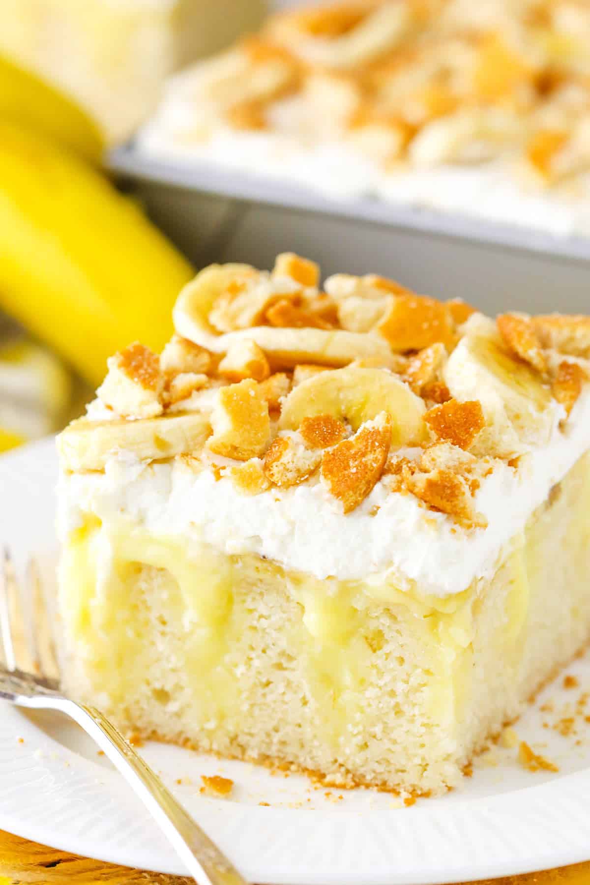 This Banana Pudding Poke Cake is made with a moist vanilla cake, homemade banana pudding, whipped cream and plenty of sliced bananas and vanilla wafers! Totally delicious and so addicting! A square serving of Banana Poke Cake next to a fork on a white plate