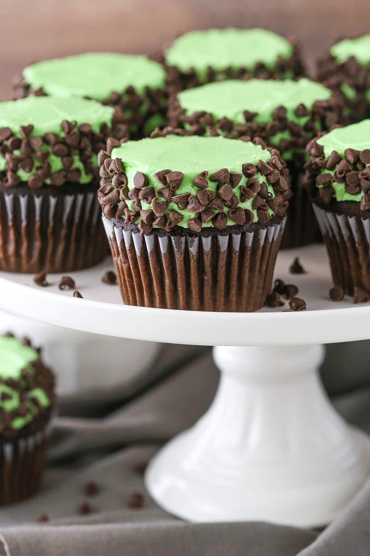 These Mint Chocolate Chip Cupcakes are made with a moist chocolate cupcake, mint frosting and mini chocolate chips! A classic flavor combination in cupcake form! Side view of Mint Chocolate Chip Cupcakes on a white cake stand
