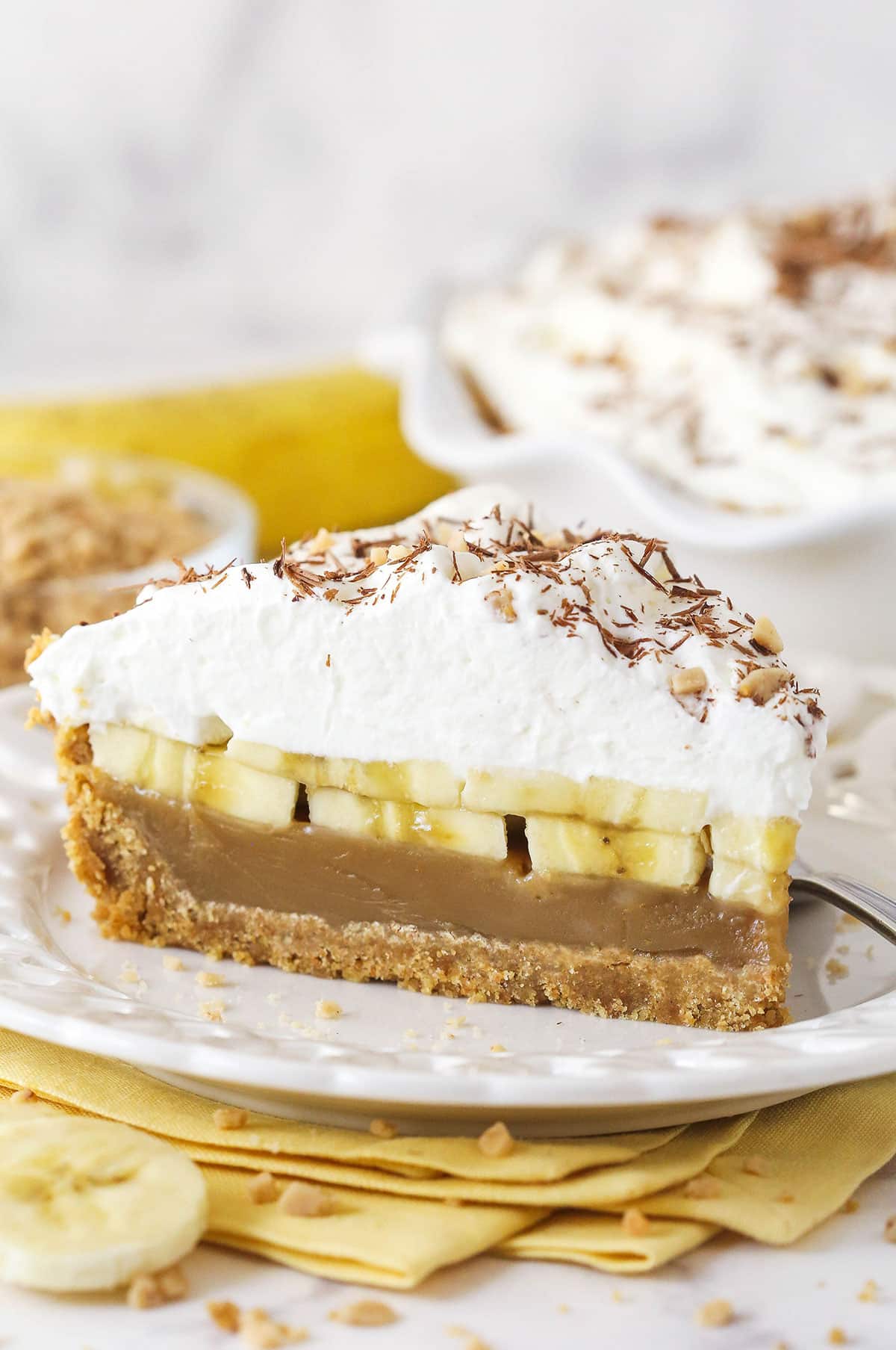Banoffee Pie is an irresistible dessert made with a graham cracker crust, toffee filling, banana slices, and whipped cream. A slice of banoffee pie on a white plate
