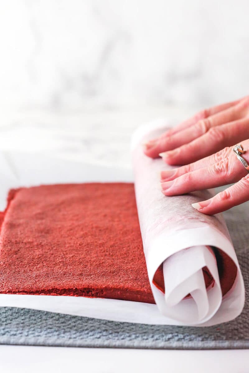 This red velvet cake roll is light and tender with the signature tang and hint of chocolate flavor. Perfect for Christmas & Valentine's Day! Two hands rolling a red velvet cake in parchment paper