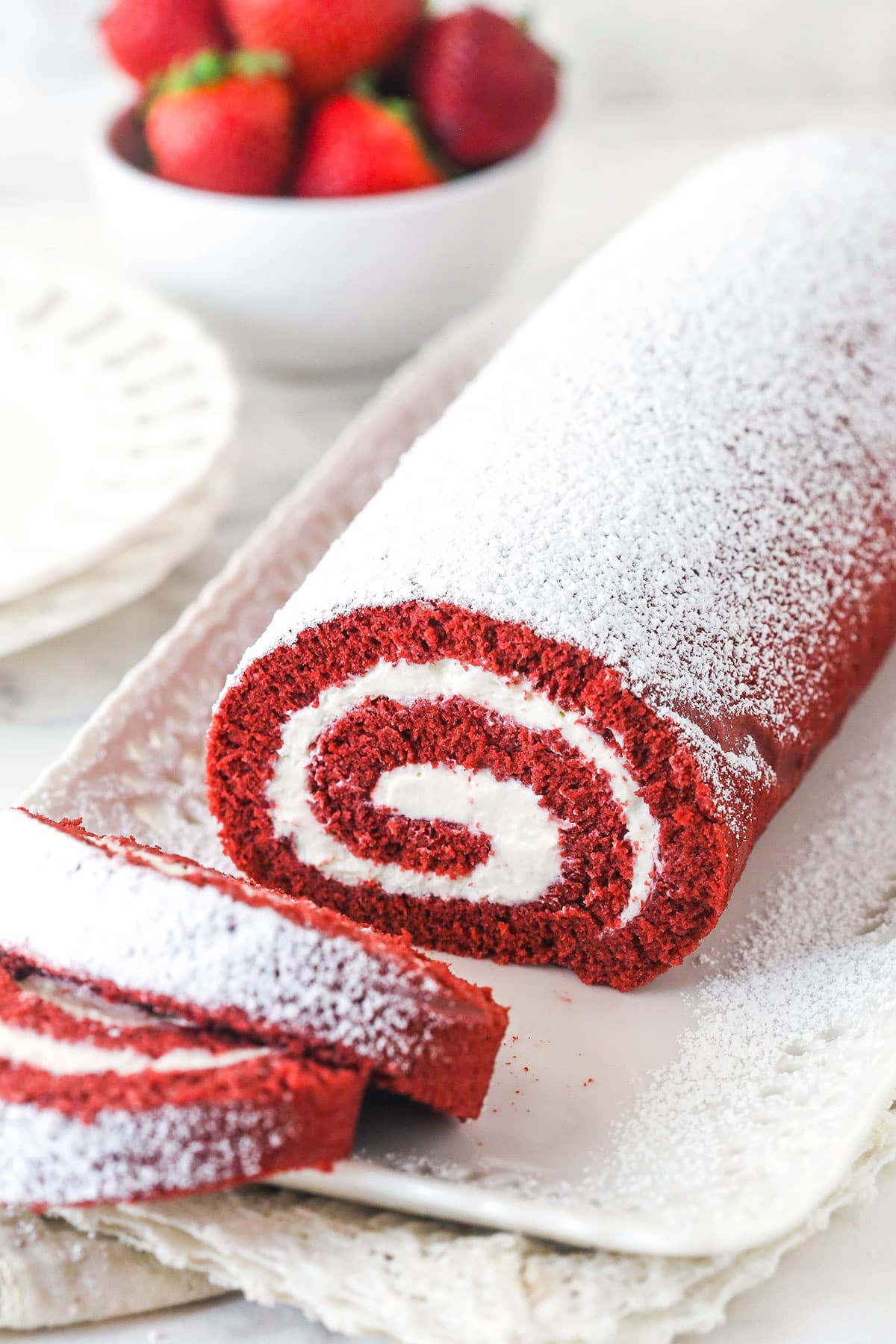 This red velvet cake roll is light and tender with the signature tang and hint of chocolate flavor. Perfect for Christmas & Valentine's Day! A red velvet cake roll on a platter with two pieces sliced