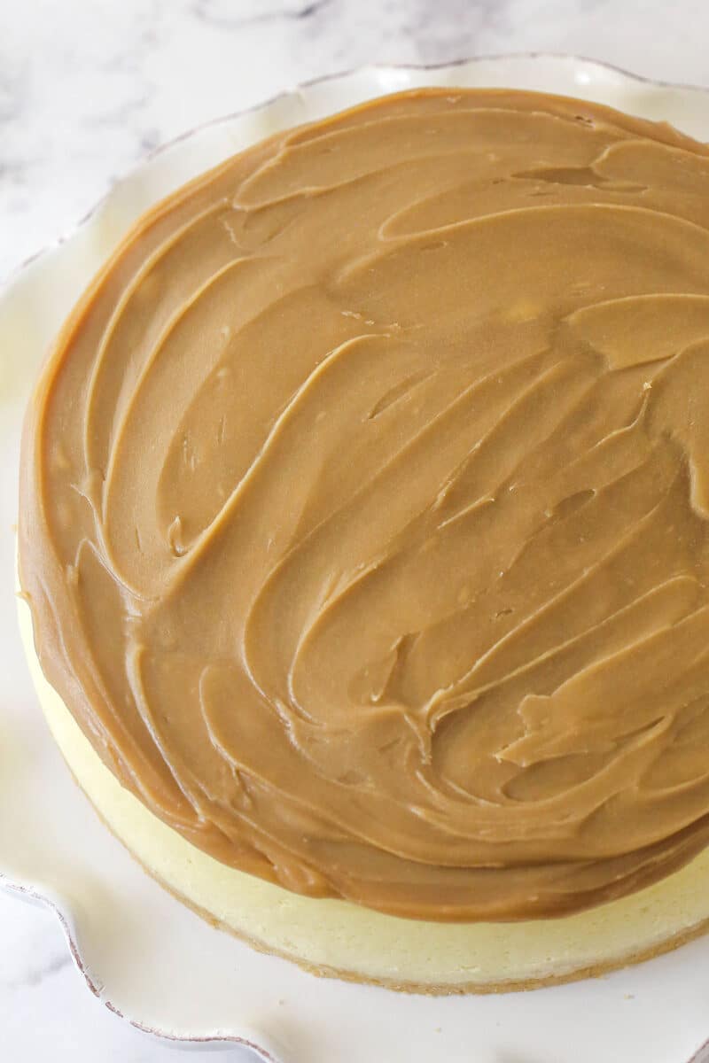 This Millionaire Cheesecake is next level! It's a tangy cheesecake doused in caramel and chocolate ganache atop a buttery shortbread crust. Spreading caramel sauce over cheesecake.