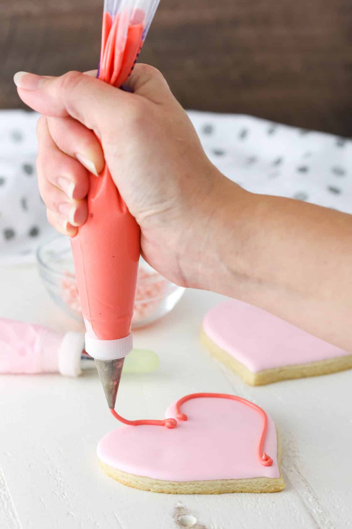 Hand piping a swirl of icing around the outside of a heart-shaped cutout cookie.