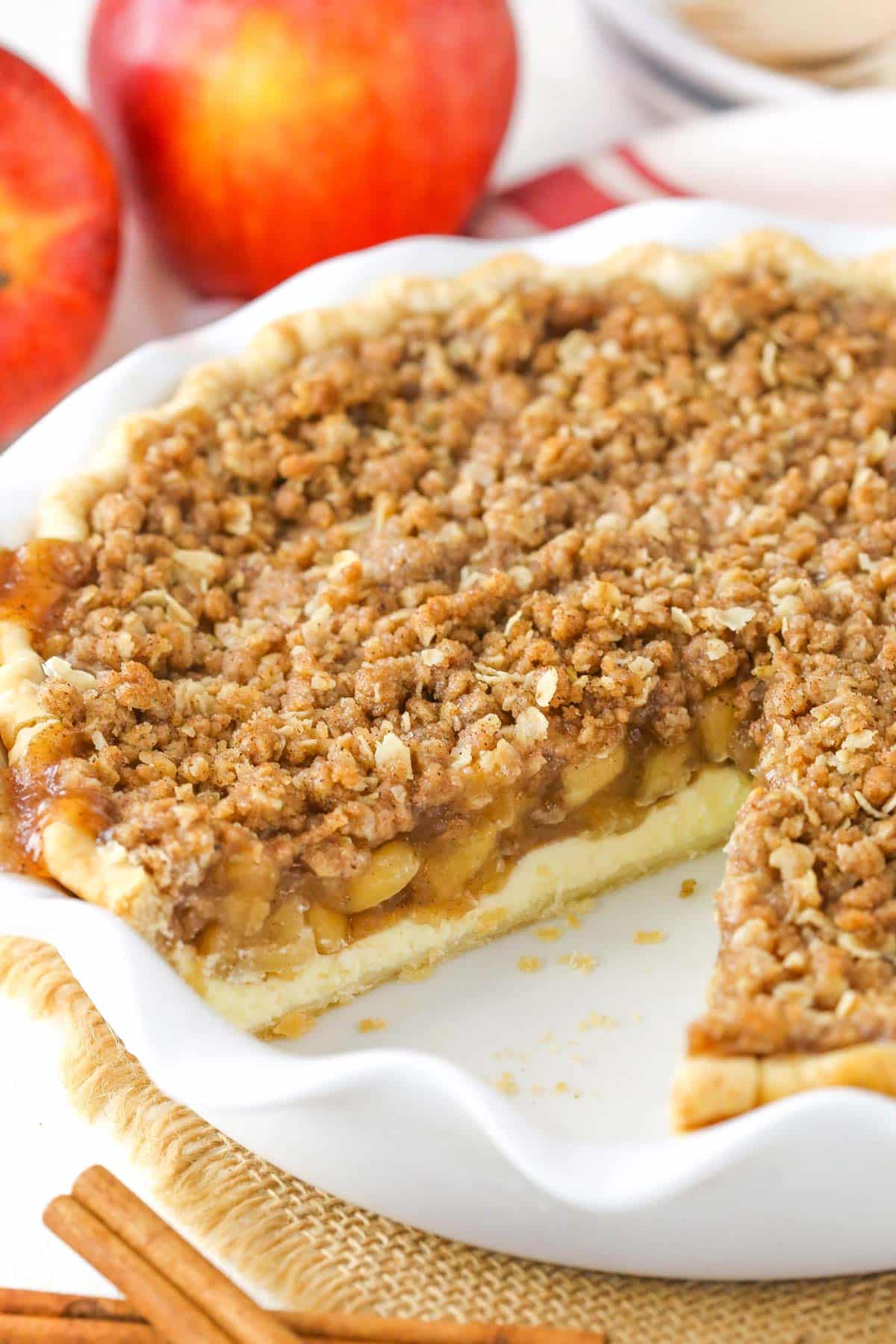 An apple crumb cheesecake pie with a slice missing in a white cake plate.