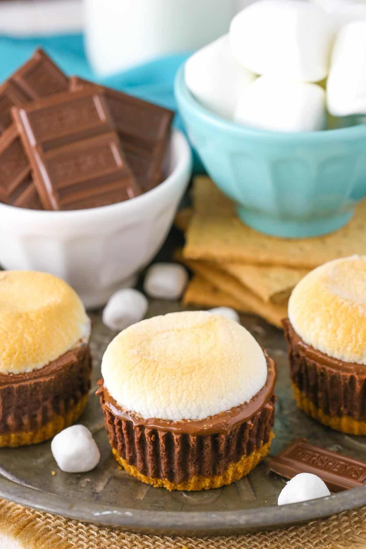 A few mini s'mores cheesecakes on a silver tray next to s'mores supplies.