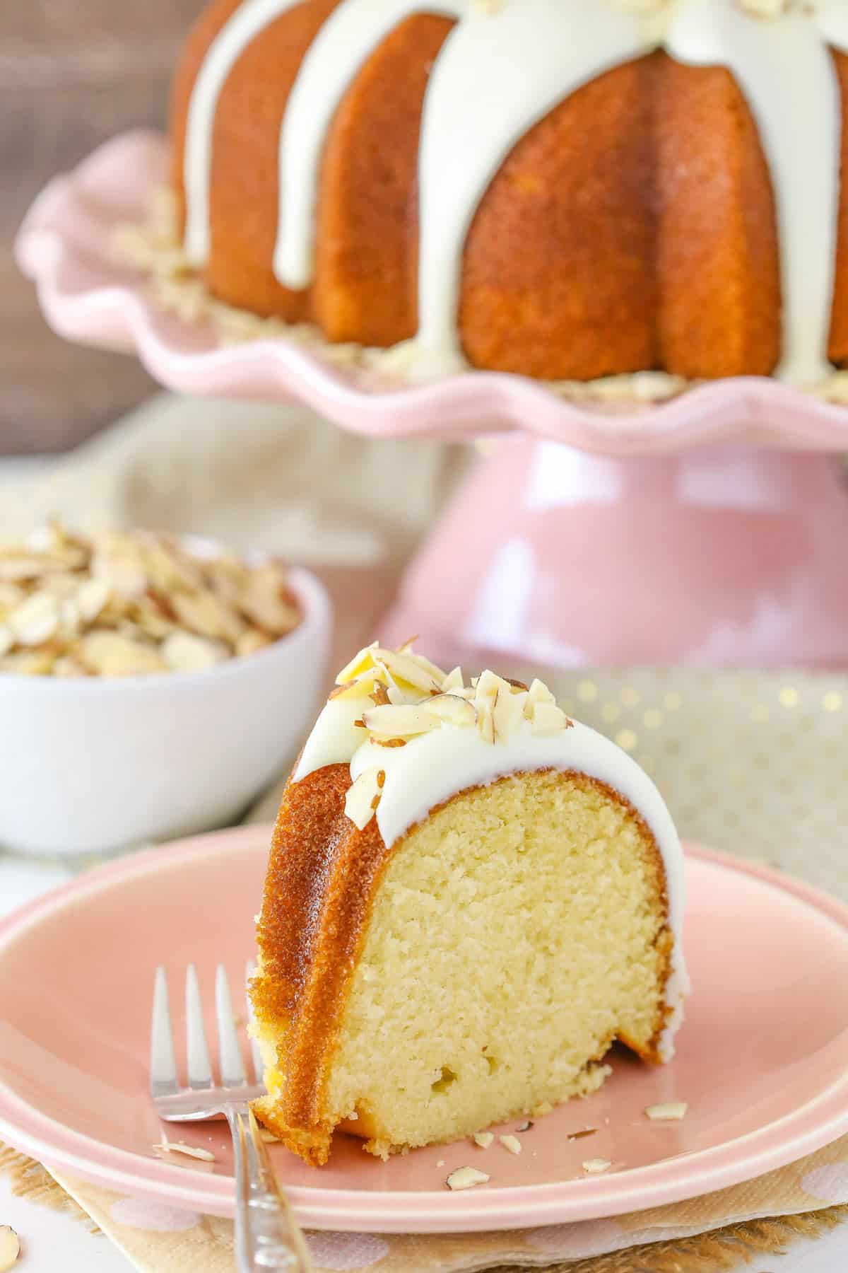 A slice of almond amaretto bundt cake on a pink plate with the rest of the cake in the background.