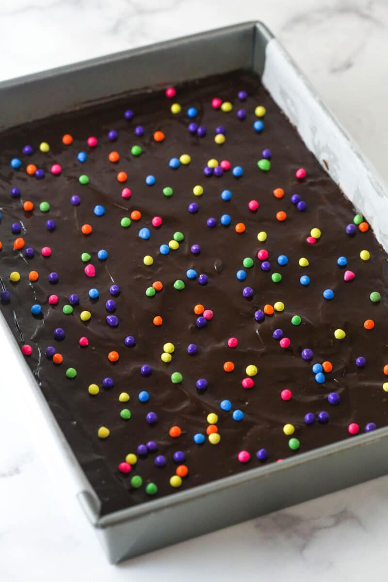 These nostalgia-filled, from-scratch Cosmic Brownies feature a fudgy brownie base topped with silky ganache and colorful sprinkles. Chocolate ganache spread over Cosmic brownies and topped with colored candy sprinkles.