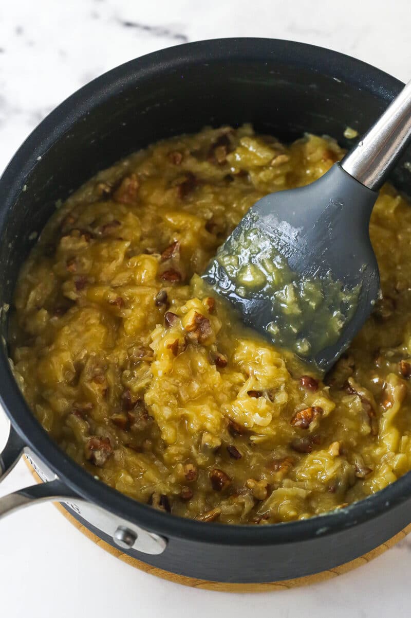 Stirring shredded coconut and toasted pecans into a thickened mixture of egg yolks, evaporated milk, vanilla, sugar, and butter.