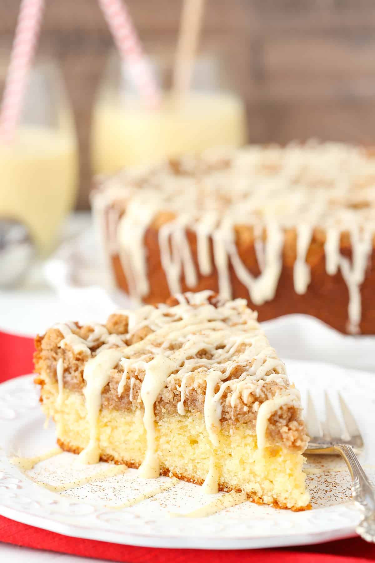 A slice of eggnog crumb cake on a white plate with the rest of the cake in the background.