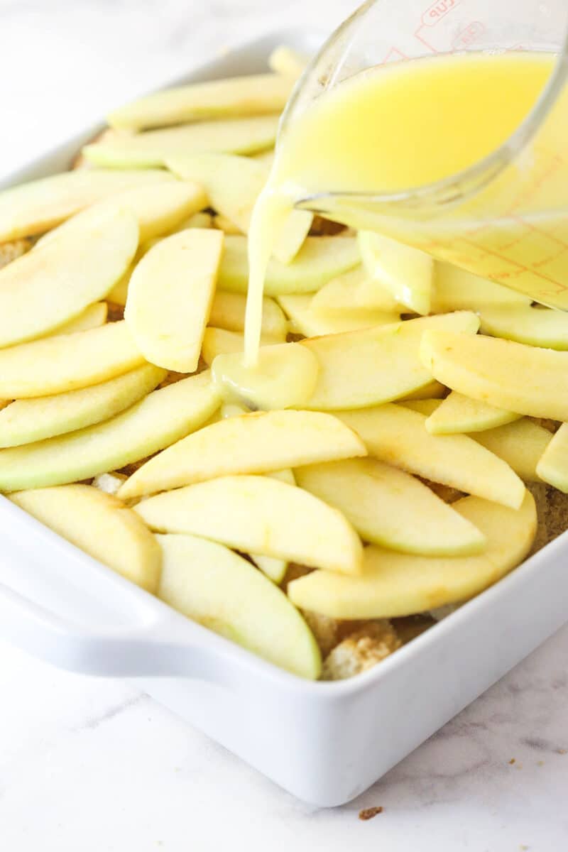 Pouring melted butter and water over apple slices that are layered with brown sugar and bread cubes.