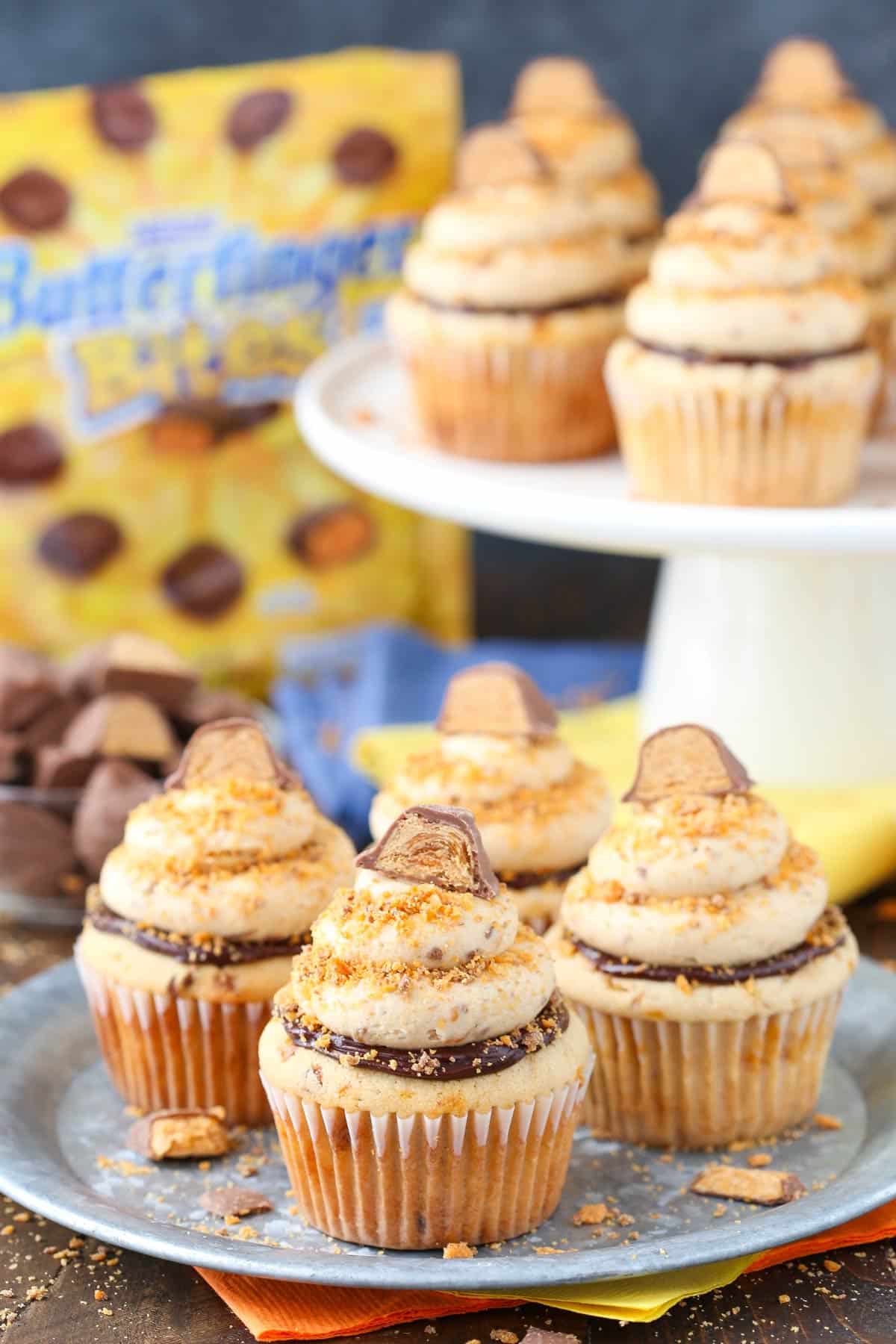 Every bite of these Butterfinger Cupcakes is loaded with crushed Butterfingers, from the moist peanut butter cupcakes to the frosting. Four Butterfinger Cupcakes on a silver plate with more cupcakes in the background.