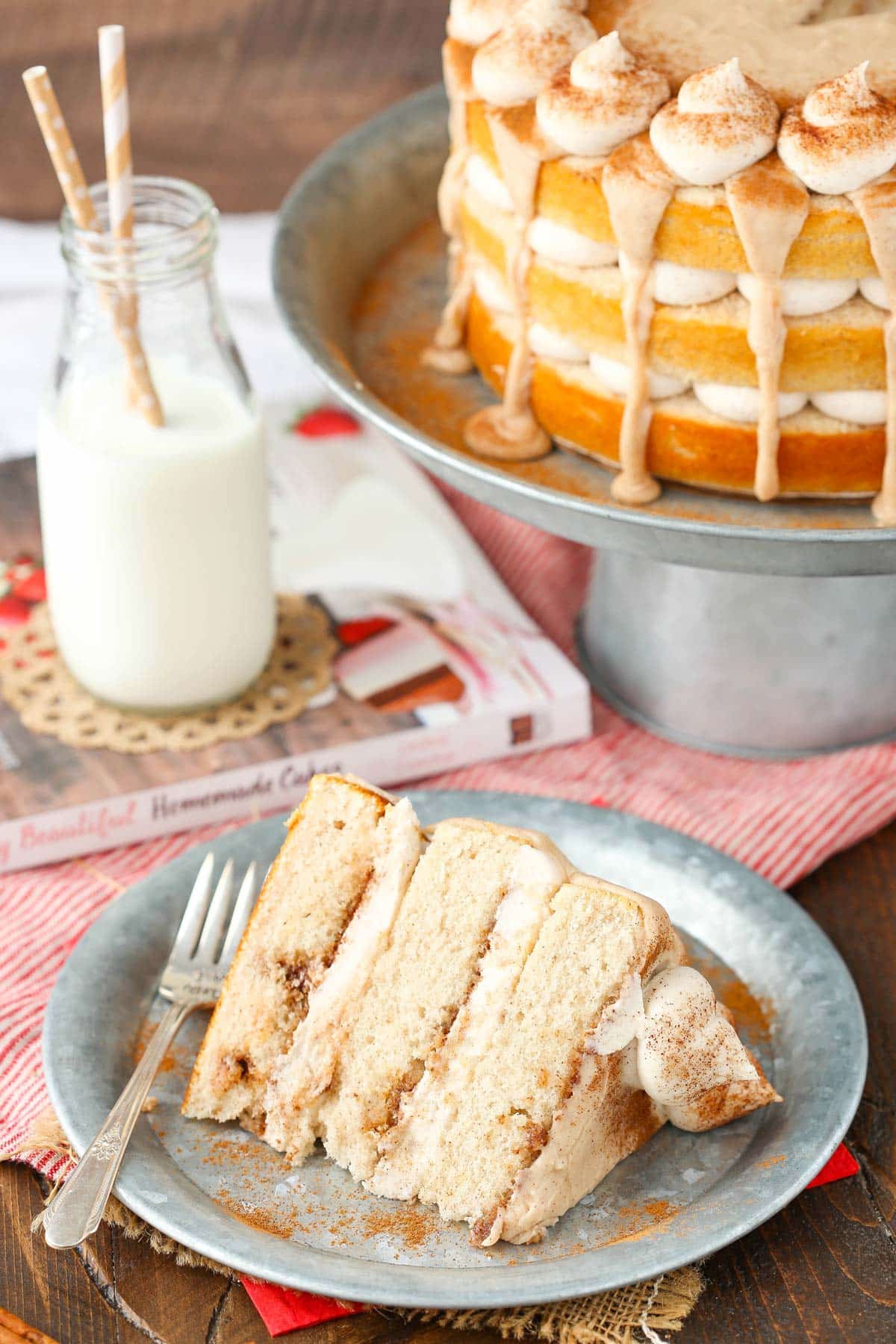A slice of cinnamon roll layer cake on a silver plate with the rest of the cake in the background.