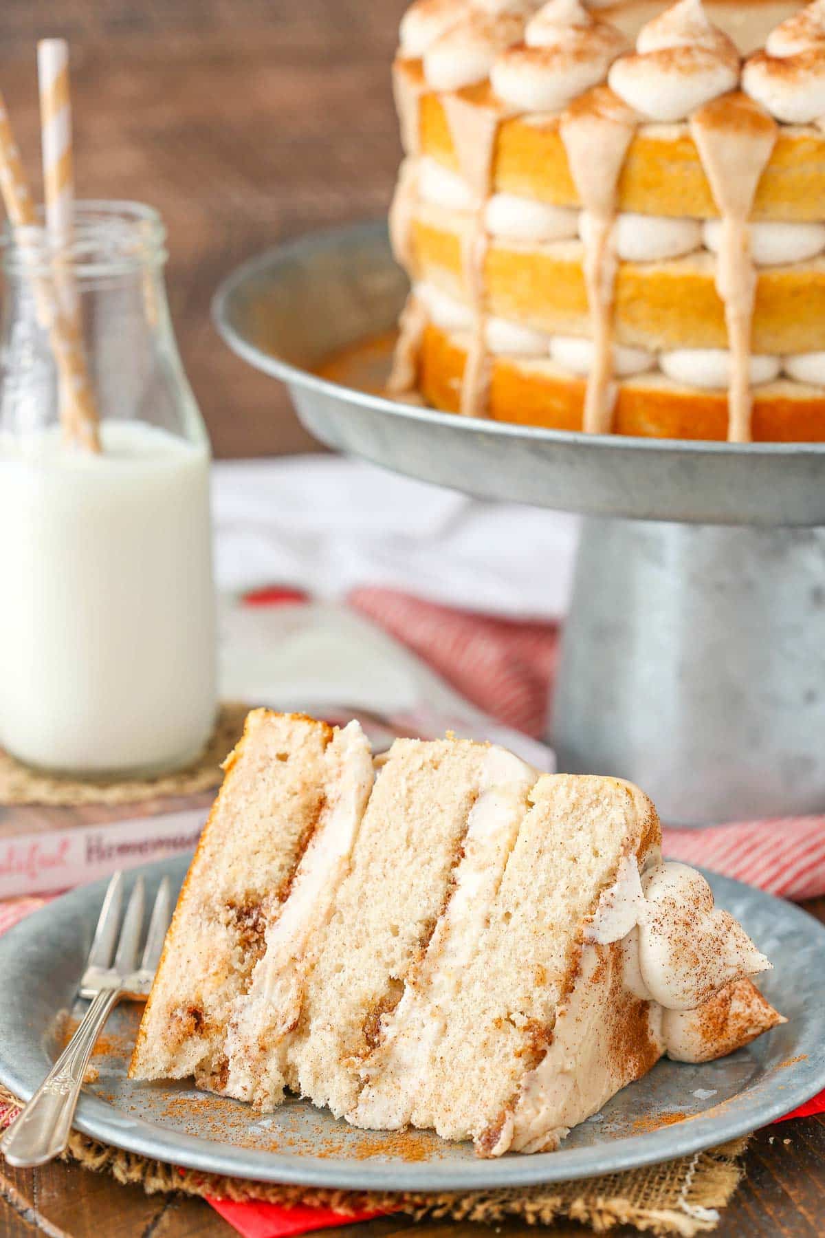 A slice of cinnamon roll layer cake on a silver plate with the rest of the cake in the background.