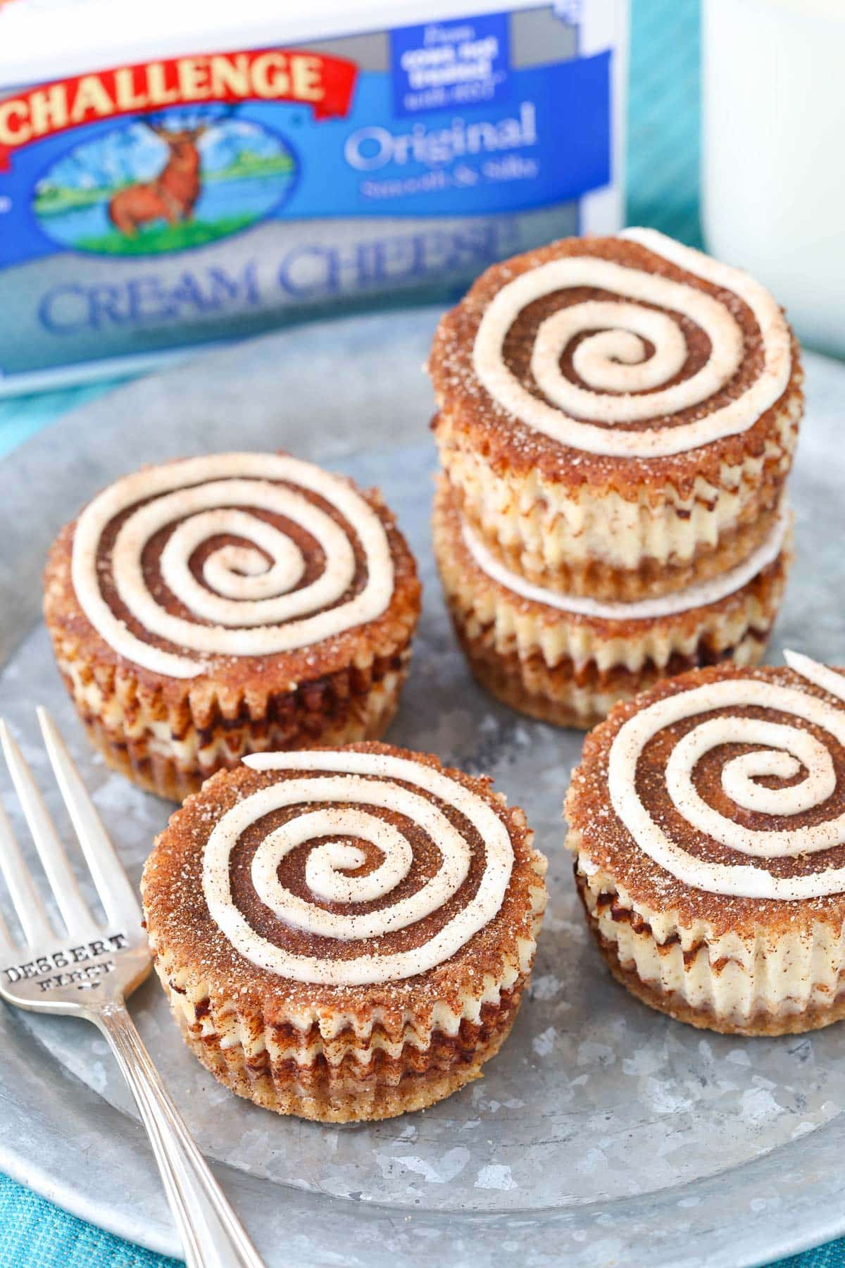 Five mini cinnamon roll cheesecakes on a silver plate next to a box of cream cheese.