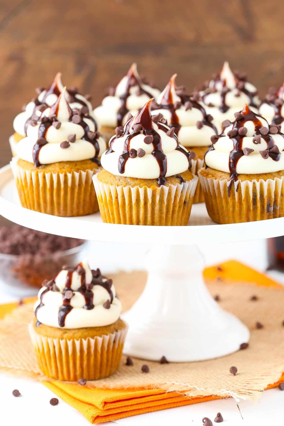 Easy pumpkin spice cupcakes filled with chocolate chips are topped with a homemade cream cheese frosting for the best fall dessert ever! A white cake stand with pumpkin chocolate chip cupcakes on top.