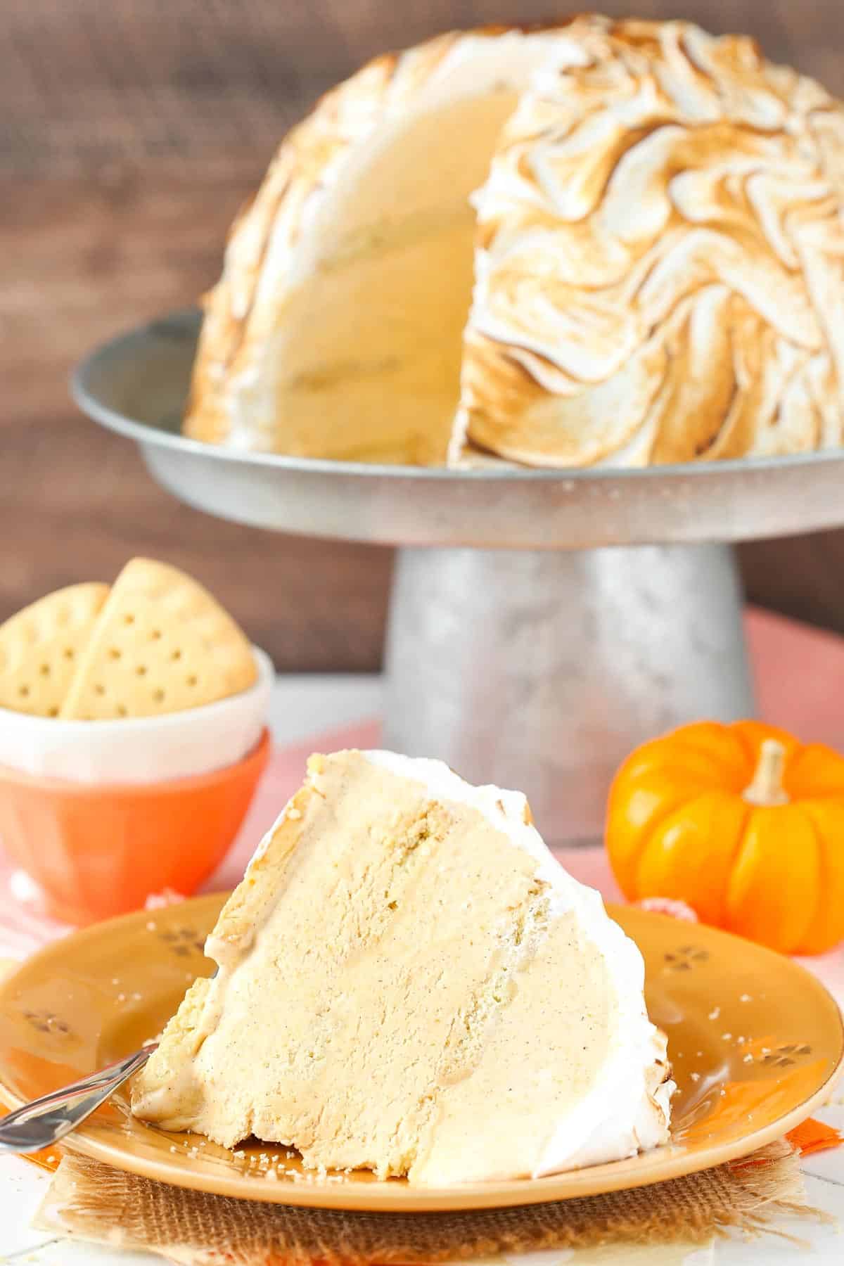 A slice of pumpkin spice baked Alaska on an orange plate with the rest of the cake in the background.