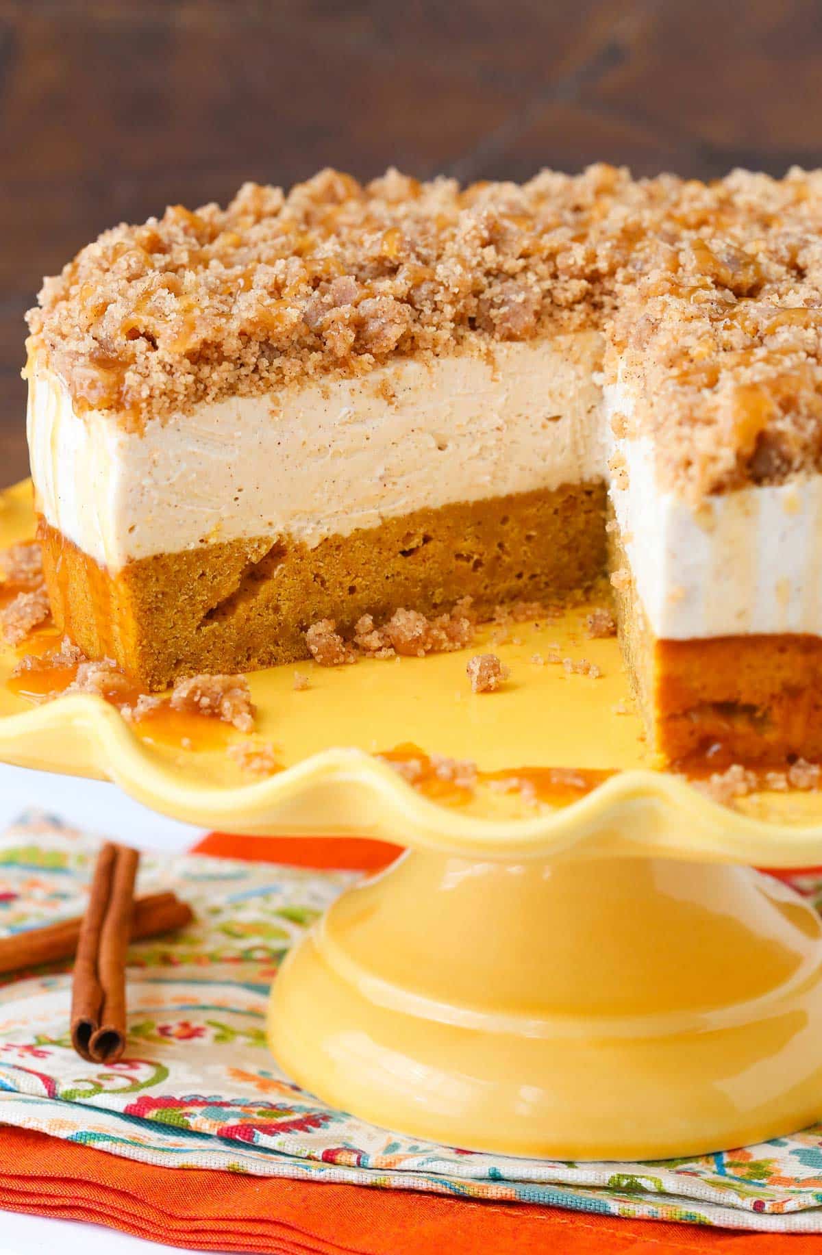 A caramel pumpkin spice blondie streusel cheesecake on a yellow cake stand with a slice removed to show layers.