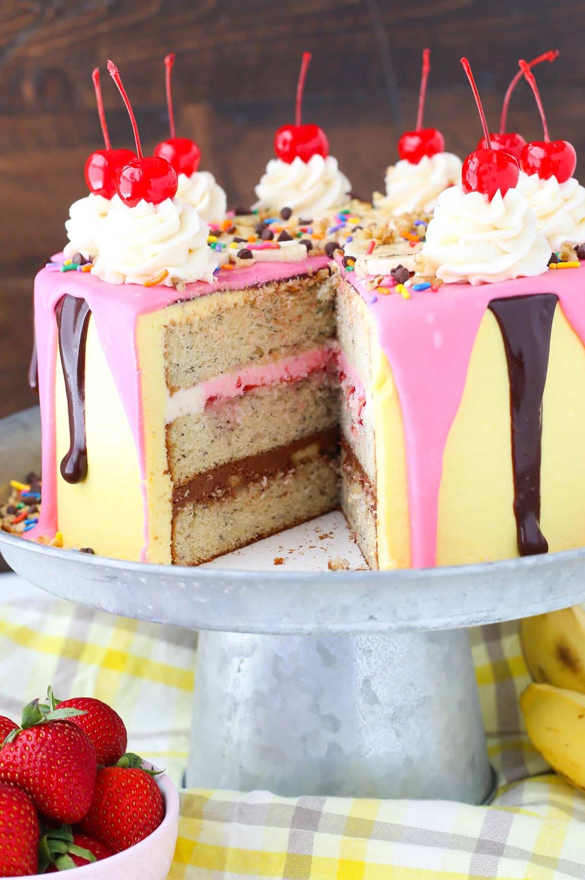 A banana split layer cake on a silver cake stand with a slice removed to show inside layers.